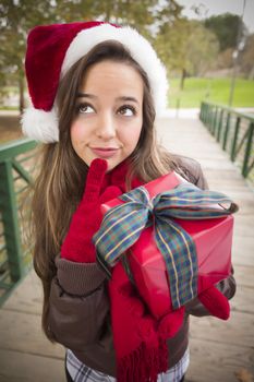 Pretty Festive Smiling Woman Wearing a Christmas Santa Hat with Wrapped Gift and Bow Outside.