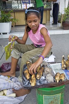 May 2012 - Bogo City, Cebu Island, Philippines - A beautiful twelve year old girl helps her mother earn money by roasting ears of corn over a charcoal fire to sell on the side of the road in Bogo City, Philippines.