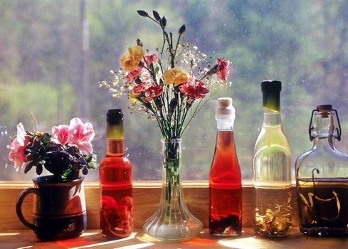 Four herb and fruit vinegar bottles on a windowsill next to two containers of flowers.