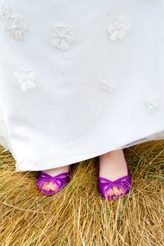 A beautiful bride in a white wedding dress stands outdoors with her pretty purple wedding shoes on her feet.