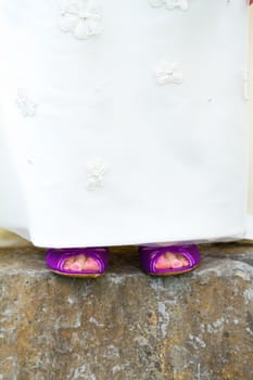 A beautiful bride in a white wedding dress stands outdoors with her pretty purple wedding shoes on her feet.