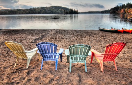Four colorful patio chairs overlooking a lake with Canoes along the shore.