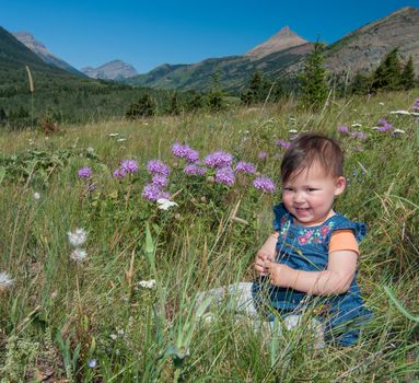 One year old baby sitting in the grass with flower around and mountains in the background.