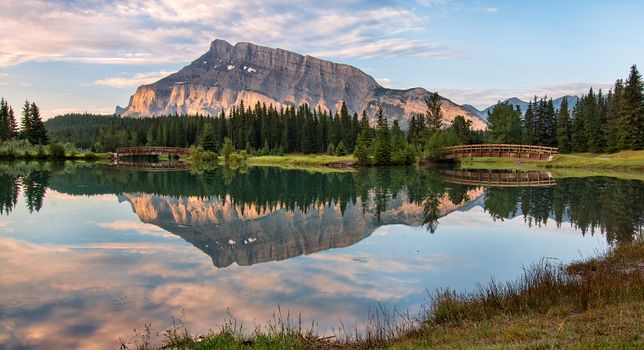 Rundle mountain reflected in pond with two bridges.