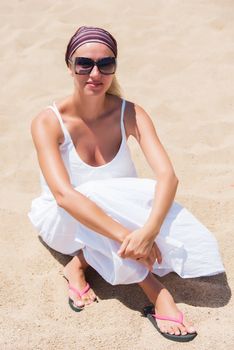 Smiling young woman in sunglasses, bandana, white dress sitting on sandy beach