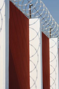 Razor wire and barbed wire on top of a red wall with white pillars, with the wire casting interesting shadows on the pillars.