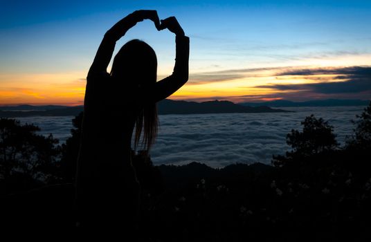 Silhouette of woman making heart shape with arms over sunset