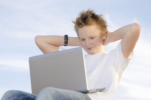 Fair-haired schoolkid leaning back with laptop against sky