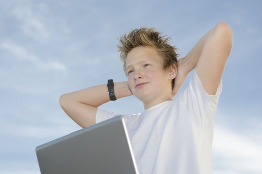 Resting teen with laptop holding hands behind his head against blue sky (high key)