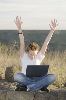 Shaggy teen with laptop joyously throws his hands up in nature