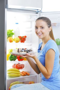 Beautiful young woman sitting near refrigerator  and choosing between healthy fruits and tasty cakes