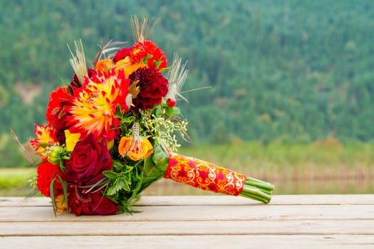 A bride's wedding bouquet sits on a wooden dock with a scenic backdrop of mountains and trees.