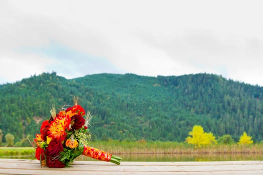 A bride's wedding bouquet sits on a wooden dock with a scenic backdrop of mountains and trees.