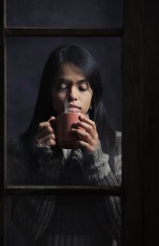 Young woman behind window with a cup of coffee or tea, focus on hands