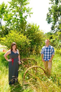 A man and a woman are in an overgrown field with rusted farm equipment.