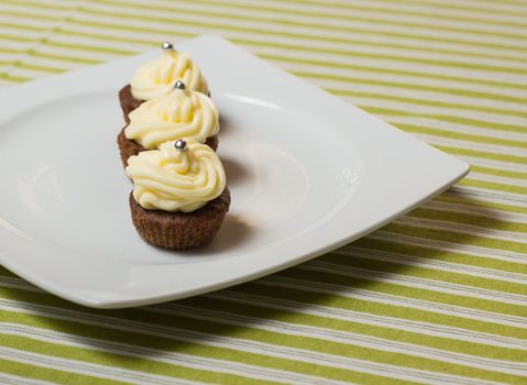 Three chocolate cupcakes with silver sprinkles on top, on white plate and fabric tablecloth with stripes