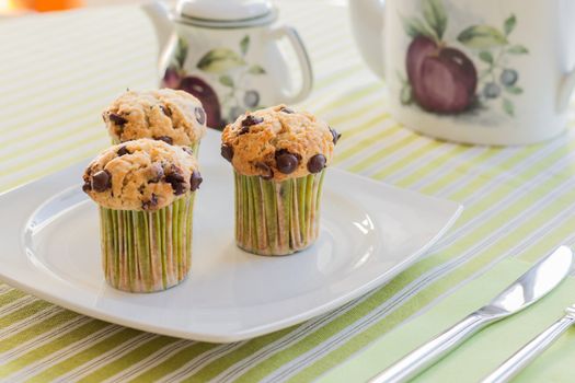 Three chocolate chip muffins on white plate and green striped tablecloth at breakfast