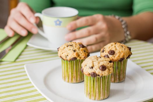 Nice girl with a cup and delicious chocolate chip muffin at breakfast in green striped tablecloth