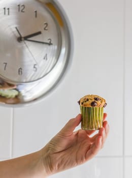 Nice girl hand taking delicious chocolate chip muffin at lunch with metalic clock in background