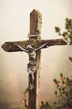 Moble photography lo-fi styled image of lichens covered figure of Christ on the Cross at the Crucifixion standing outdoors amonst pine branches against a misty fall forest backdrop