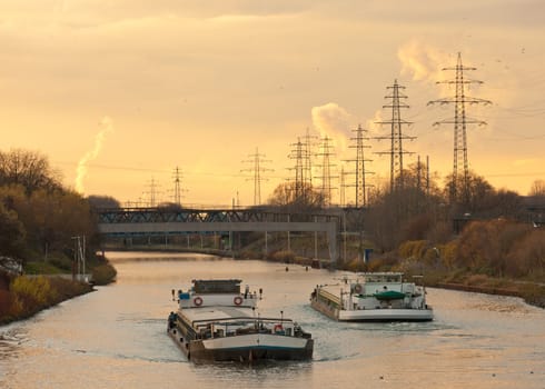 Inland waterway vessels on a busy channel in industrial area near a bridge under the warm glow of an evening sunset