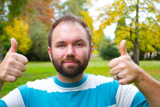 A man with a full beard gives the camera a double thumgs up with both hands while outdoors in a park setting.