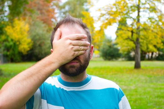 A man with a full beard covers his eyes and face while outdoors in a park setting.