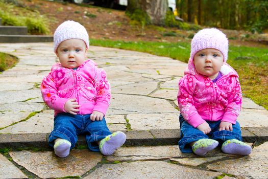 Two twin baby girls sit on a stone walkway wearing pink sweatshirts and beanies.