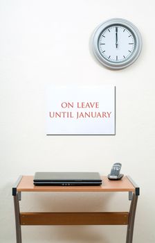 On leave until January sign, closed laptop, office desk, clock on wall