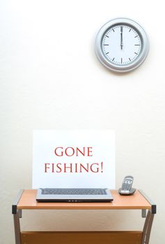 Office desk with gone fishing sign on open laptop next to portable phone, clock on wall