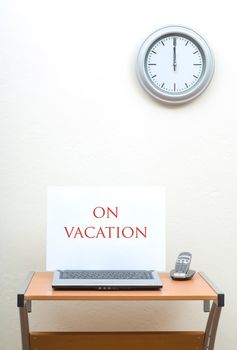 Laptop with on vacation sign next to portable phone on top of office desk, clock on wall