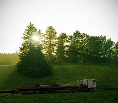 Sun and forest trees shine down on long semi truck delivery with motion blur on highway road