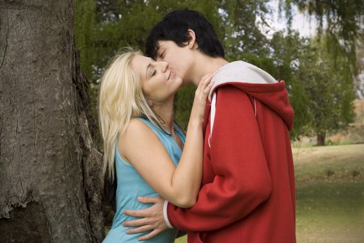 Boyfriend with red jacket kissing girlfriend next to tree in garden or park