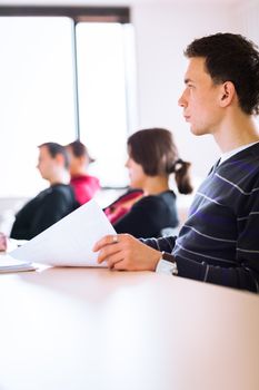 young, handsome male college student sitting in a classroom full of students during class (color toned image; shallow DOF)