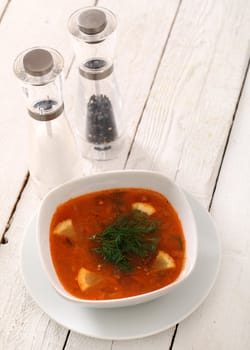 Image of bowl of hot red soup served with pepper, salt and spoon on white wooden table