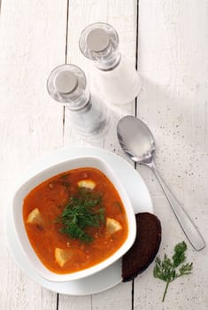 Image of bowl of hot red soup served with bead, pepper, salt and spoon on white wooden table