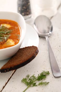 Image of bowl of hot red soup served with bead, pepper, salt and spoon on white wooden table