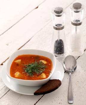 Image of bowl of hot red soup served with bead, pepper, salt and spoon on white wooden table