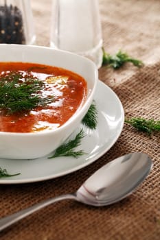 Image of bowl of hot red soup served with the salt, pepper and spoon on a beige tablecloth