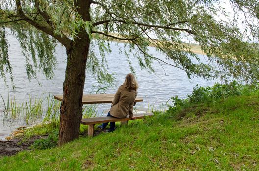 young blond woman sit on wooden bench under willow tree branches and admire lake landscape.