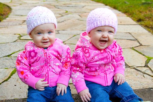 Two twin baby girls sit on a stone walkway wearing pink sweatshirts and beanies.