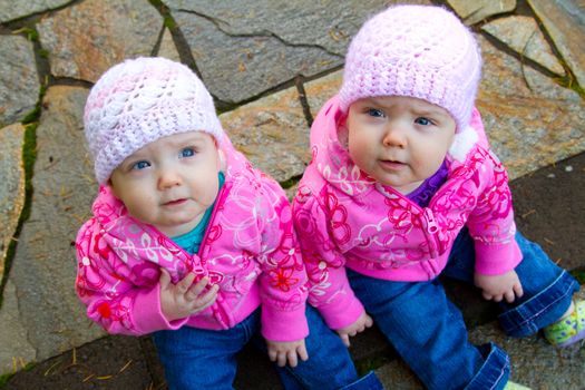 Two twin baby girls sit on a stone walkway wearing pink sweatshirts and beanies.