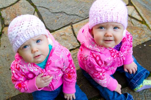 Two twin baby girls sit on a stone walkway wearing pink sweatshirts and beanies.