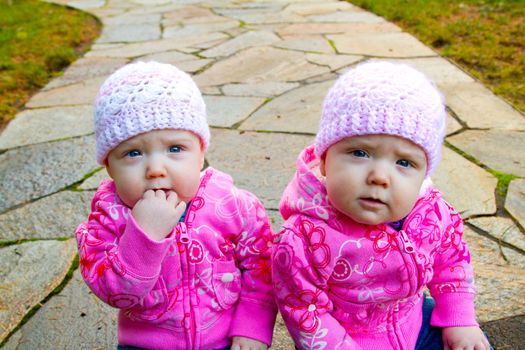 Two twin baby girls sit on a stone walkway wearing pink sweatshirts and beanies.