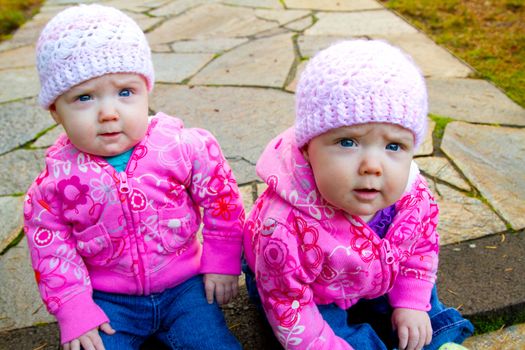 Two twin baby girls sit on a stone walkway wearing pink sweatshirts and beanies.