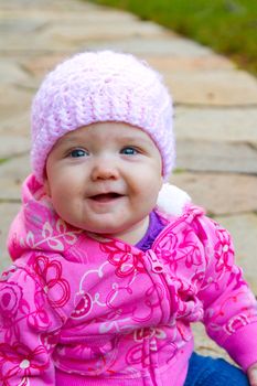 A young infant girl wearing a pink sweatshirt and a knitted cap sits on a stone walkway for a portrait.