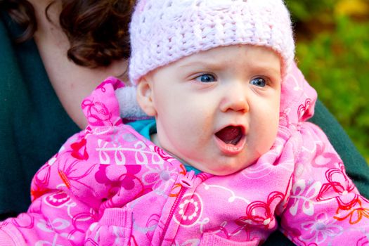 A young infant child yells with her mouth open while her mom holds her outdoors.