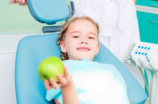 girl in the dentist's chair shows a green apple, regular care of your teeth