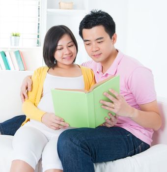Asian couple reading book together, sitting on sofa