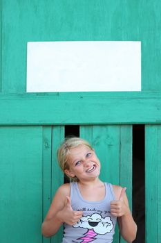 A young girl doing thumbs up and pulling a funny face under an empty sign. Space for text